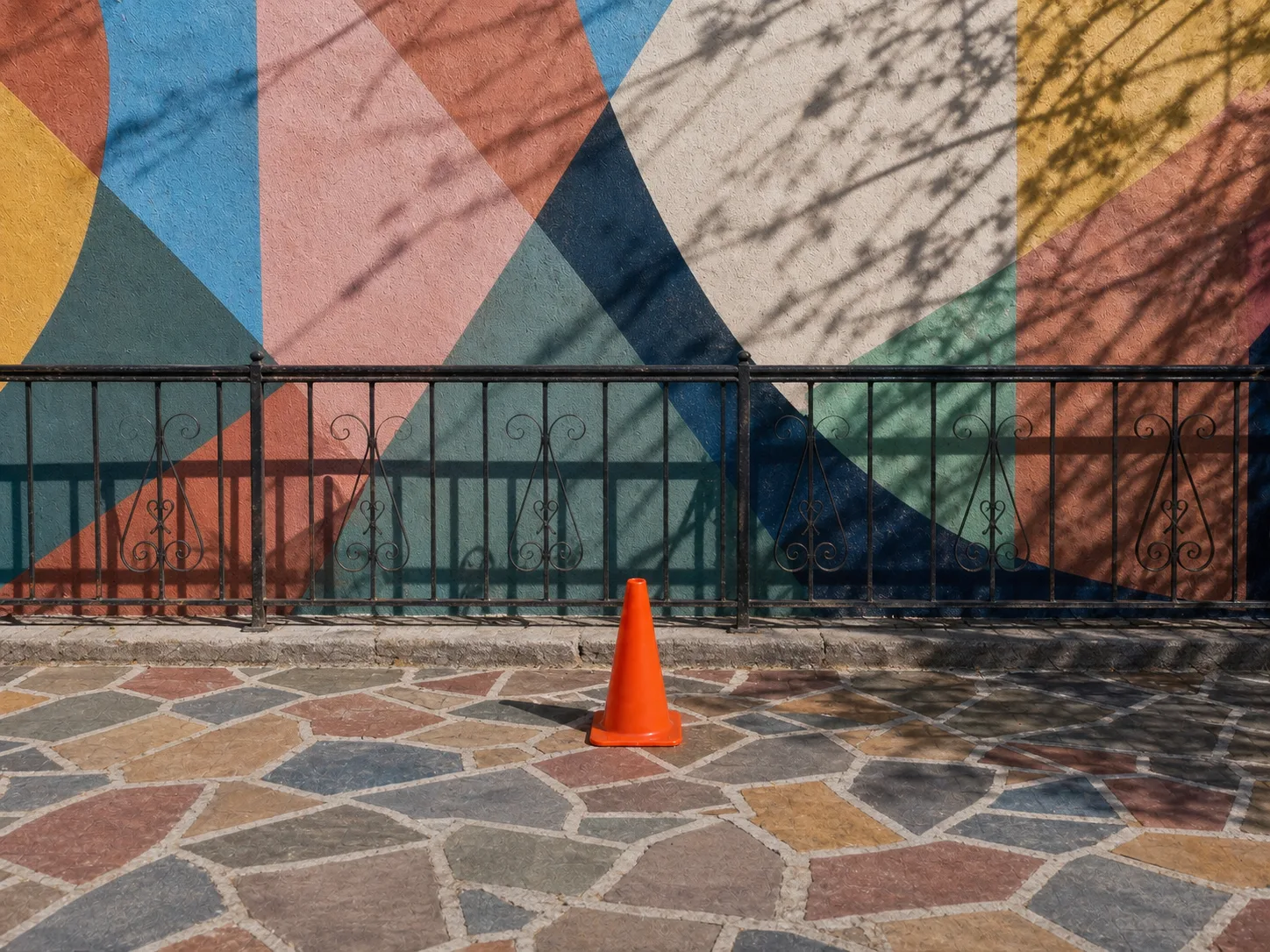 Avoid example of an orange cone against a geometric mural, iron railing, mosaic paving, and crossing shadows